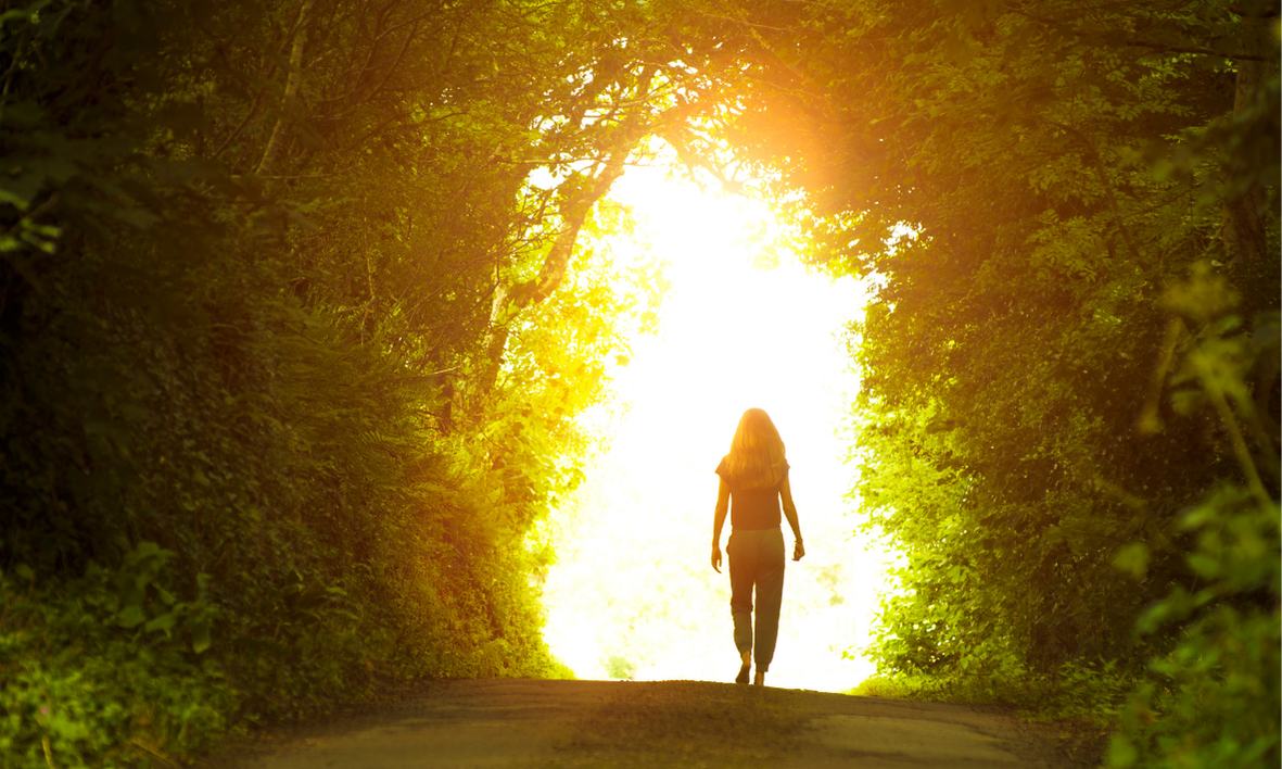 Woman walks toward brilliant sunlight through leafy green trees, emerging into the light.