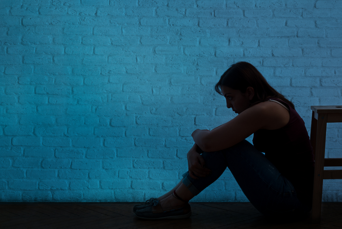 Woman sits on the floor, alone in a blue-lit room, hugging her knees.
