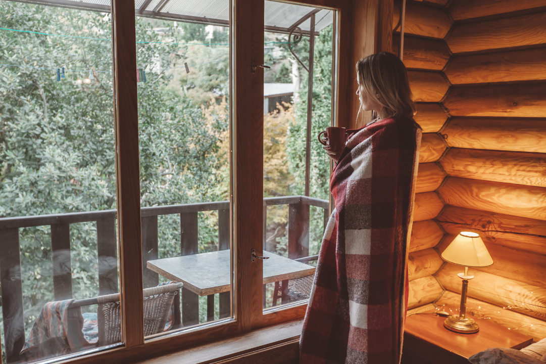 A woman in a blanket holding coffee, staring out a cabin window, surrounded by autumn forest