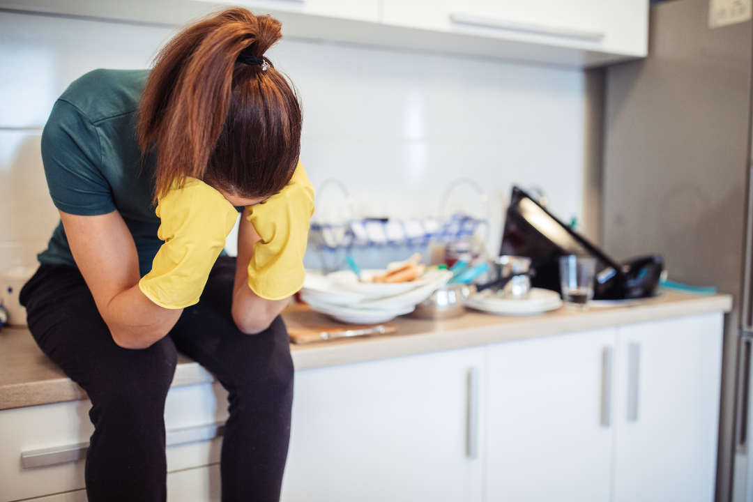 Exhausted woman sitting on a kitchen counter with rubber gloves, head in hands, surrounded by household clutter, representing emotional and physical burnout.