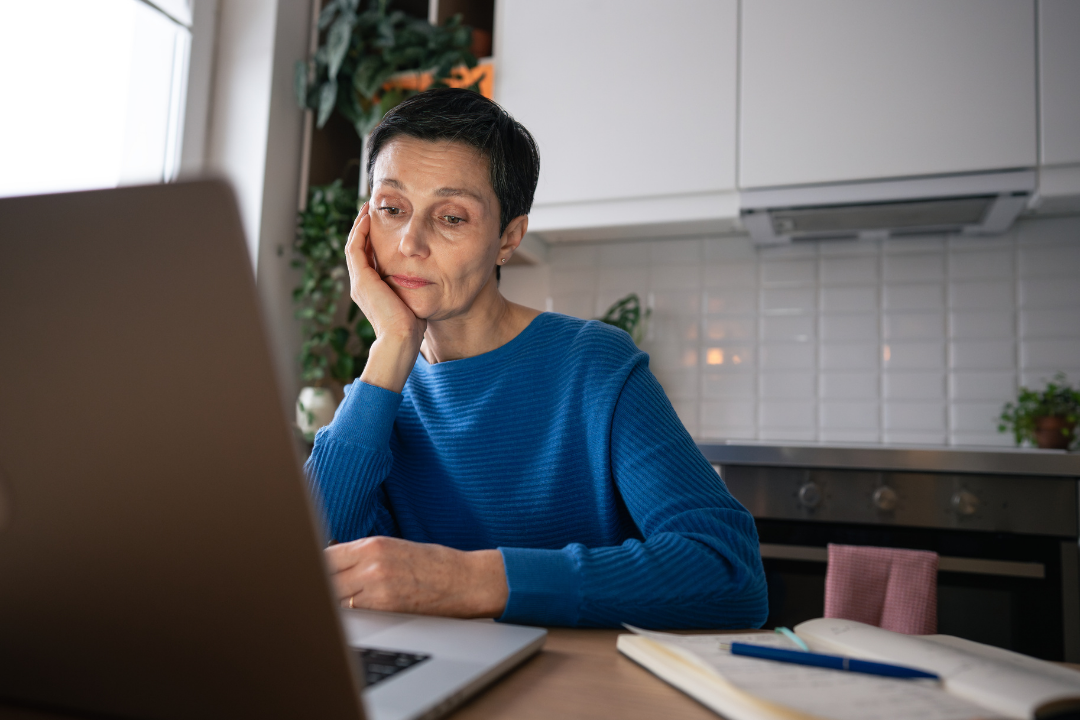 Woman in a blue sweater staring tiredly at a laptop, sitting at a kitchen table